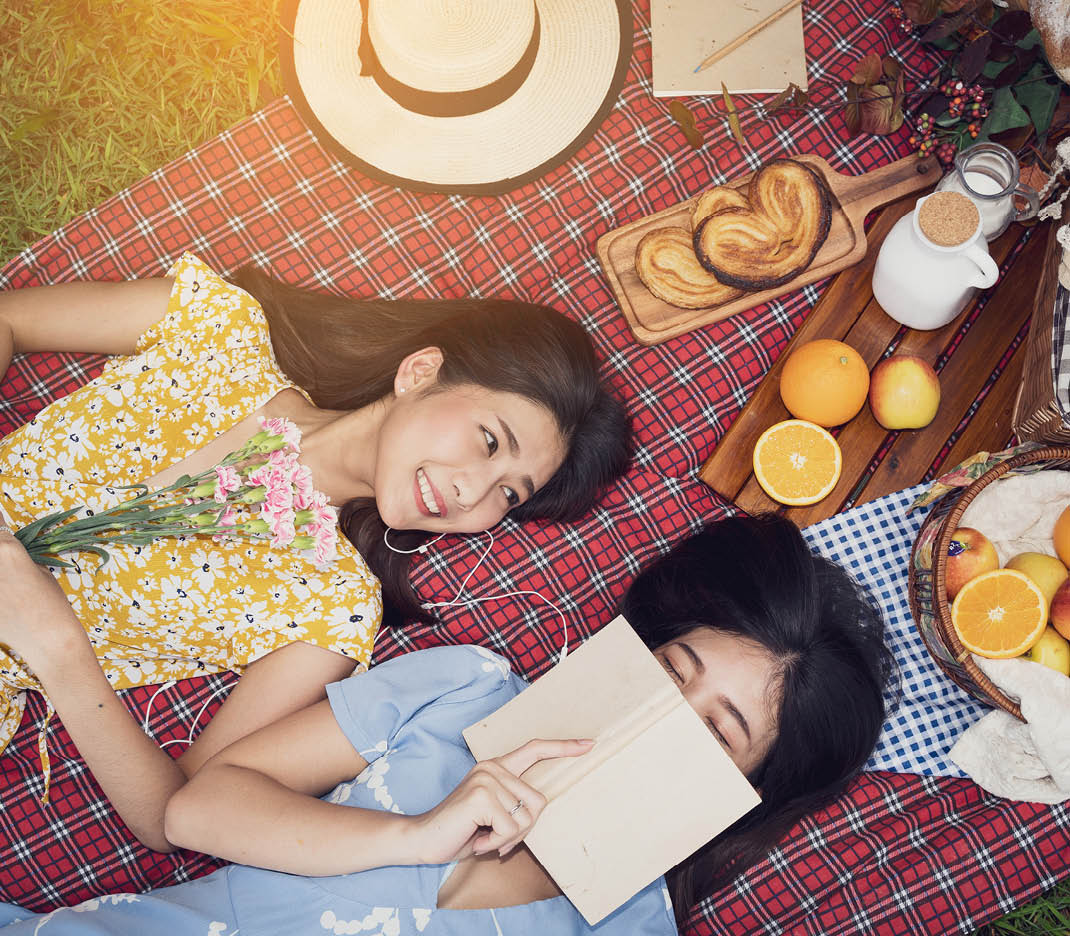 Couple of girls going picnic in a park, reading a book together and feeling friendly.